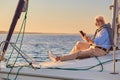 Side view of a retired man sitting on the side of his sailboat or yacht floating in the calm blue sea at sunset, using