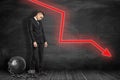 Side view of sad businessman standing shackled to big metal ball and looking down near black graph-ruled wall with red Royalty Free Stock Photo