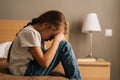 Side view of sad little girl hugging knee, sobbing with head bowed and crying sitting alone on bed in bedroom. Royalty Free Stock Photo