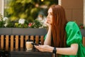 Side view of sad redhead young woman holding mobile phone, sitting at table with coffee cup in outdoor cafe terrace in Royalty Free Stock Photo