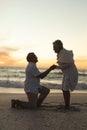 Side view of senior biracial man proposing woman while kneeling on shore at beach during sunset Royalty Free Stock Photo