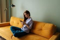 Side view of serious redhead young woman looking at smartphone screen sitting on yellow sofa, chatting online, using Royalty Free Stock Photo