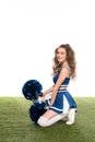 View of sexy happy cheerleader girl in blue uniform with pompoms sitting on grassy field isolated on white