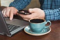 Side view shot of a man`s hands using smart phone and laptop sitting at wooden table with cup of black coffee. Close up. Royalty Free Stock Photo