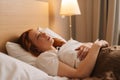 Side view of sick exhausted young woman with flu lying under blanket on bed holding thermometer in armpit, looking away Royalty Free Stock Photo