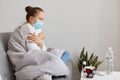 Side view of sick woman has influenza, catching cold or coronavirus, sitting on sofa wrapped in blanket and wearing medical mask, Royalty Free Stock Photo