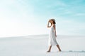 View of smiling beautiful girl in white dress and straw hat walking on sandy beach with blue sky Royalty Free Stock Photo