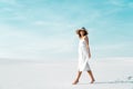 View of smiling beautiful girl in white dress and straw hat walking on sandy beach with blue sky Royalty Free Stock Photo