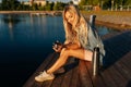 Side view of smiling blonde young woman holding cup with hot coffee from thermos sitting on beach by wooden pier on Royalty Free Stock Photo