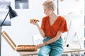 side view of smiling female freelancer sitting on table and eating pizza at home