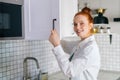 Side view of smiling redhead young woman opening door of kitchen cabinet at light kitchen room. Royalty Free Stock Photo