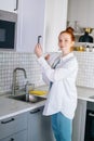 Side view of smiling redhead young woman opening door of kitchen cabinet at light kitchen room. Royalty Free Stock Photo