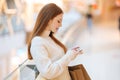 Side view of smiling redhead young woman using smartphone holding shopping paper bags with purchase standing in hall of Royalty Free Stock Photo