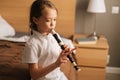 Side view of talented adorable little girl playing flute sitting on bed in bedroom, looking away. Royalty Free Stock Photo