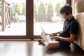 Side view of teenager boy wearing black headphones, sitting on floor, putting laptop on legs, typing, surfing internet.