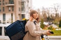 Side view of thinking food delivery woman with thermo backpack holding mobile phone waiting next order standing with