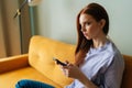 Side view of thoughtful beautiful redhead woman holding cellphone, sitting on cozy home sofa, looking away, thinking Royalty Free Stock Photo