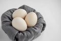 Side view of three unpainted beige eggs in the nest of the grey fabric on a white background.