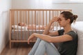 Side view of mother with postnatal depression symptoms sitting on floor while baby sleeping in bed