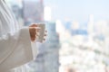 Side view of woman in bathrobe standing near the window while holding white cup of coffee and enjoy wonderful morning in the hotel Royalty Free Stock Photo