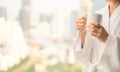 Side view of woman in bathrobe standing near the window while holding white cup of coffee and enjoy wonderful morning in the hotel Royalty Free Stock Photo