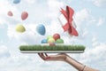 Side view of woman`s hand holding tray owith patch of green lawn with open gift box on top from which colorful balloons