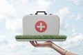 Side view of woman`s hand holding tray with patch of green grass on it with doctor`s case on top against cloudy sky. Royalty Free Stock Photo