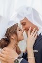 Side view of smiling couple standing under bridal veil. Young woman touching face, young handsome groom hugging bride. Royalty Free Stock Photo