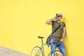 Side view of a young adult man with a vintage bike and wearing casual clothes and sunglasses standing against a yellow wall while Royalty Free Stock Photo