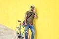Side view of a young adult man with a vintage bike and wearing casual clothes and sunglasses standing against a yellow wall while Royalty Free Stock Photo