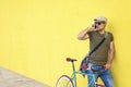 Side view of a young adult man with a vintage bike and wearing casual clothes and sunglasses standing against a yellow wall while Royalty Free Stock Photo