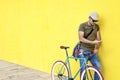Side view of a young adult man with a vintage bike and wearing casual clothes and sunglasses standing against a yellow wall while Royalty Free Stock Photo