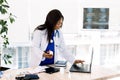 Side view of young African-american female doctor holding a bottle of medicine and using laptop while working at desk in Royalty Free Stock Photo