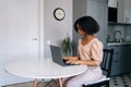 Side view of young African woman sitting at table in modern kitchen room working on laptop looking to screen. Pretty Royalty Free Stock Photo