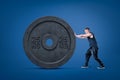 Side view of young athletic little man in sleeveless hoody pushing huge black 25kg weight plate on navy blue background.