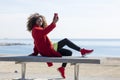 Side view of young beautiful curly african american woman sitting on a bench at beach while using a mobile phone outdoors Royalty Free Stock Photo