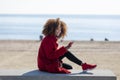 Side view of young beautiful curly african american woman sitting on a bench at beach while using a mobile phone outdoors Royalty Free Stock Photo