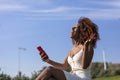 Side view of a young beautiful curly afro woman sitting on ground in a park and using a mobile phone while smiling in a sunny day Royalty Free Stock Photo