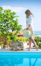 Side view of young beautiful woman in white shirt and hat standing near blue water swimming pool on blur landscape background Royalty Free Stock Photo