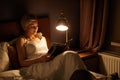 Side view of young caucasian woman sitting on bed reading favourite interesting book at home in room lighted by lamp