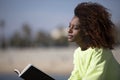 Side view of a young curly afro woman sitting on a breakwater holding a book while reading outdoors Royalty Free Stock Photo