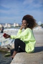 Side view of a young curly afro woman sitting on breakwater rocks enjoying and smiling while using a mobile phone to listen music Royalty Free Stock Photo