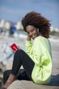 Side view of a young curly afro woman sitting on breakwater rocks enjoying and smiling while using a mobile phone to listen music Royalty Free Stock Photo