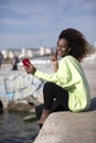 Side view of young curly afro woman sitting on breakwater rocks enjoying and smiling while using a mobile phone to listen music in Royalty Free Stock Photo