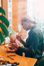 Side view of young female master reborn maker decorating newborn caucasian baby doll while sitting in home workshop. Royalty Free Stock Photo