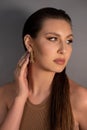 Side view of young mysterious woman with long dark hair, shining make-up, wearing golden earrings, ring, touching neck.