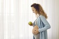 Side view of a young pregnant woman standing by the window and eating a fresh apple Royalty Free Stock Photo