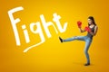 Side view of young pretty girl in jeans, sleeveless top and red boxing gloves with leg lifted for a kick on yellow Royalty Free Stock Photo