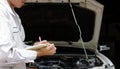 Side view of young professional mechanic in uniform writing on clipboard against car in open hood at the repair garage. Insurance Royalty Free Stock Photo