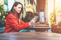 Side view.Young smiling attractive woman in orange coat is sitting outside in cafe at table and uses tablet computer. Royalty Free Stock Photo